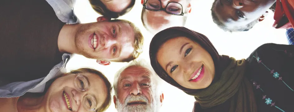 A group of seven people of different ages, genders and ethnicities stand with their heads close together, smiling down at the camera.